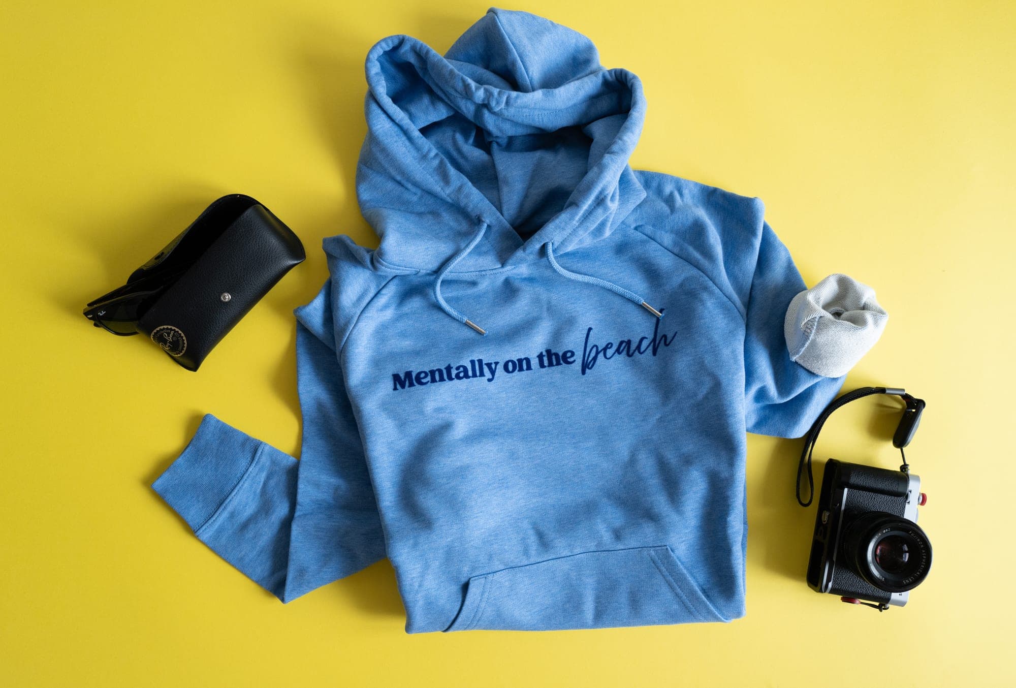 Sky blue hoodie with “Mentally on the beach” in navy, laid flat on yellow with sunglasses and a camera